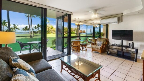A modern living room with a sofa, a glass coffee table, and a television. There is a balcony with a view of palm trees and a golf course. - Kihei Surfside, #111 1 Bedroom Condo by RedAwning (Kihei)
