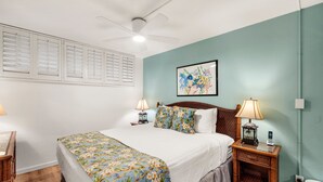 A hotel room with a bed, two bedside tables, a ceiling fan, a wall-mounted picture, and a window with blinds. - Kihei Surfside, #508 1 Bedroom Condo by RedAwning (Kihei)