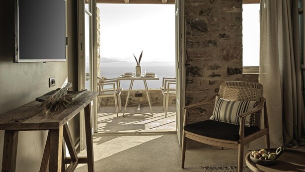 Une pièce avec un mur en pierre, une table en bois ornée d’un vase et de verres, une chaise en osier et une vue sur la mer.