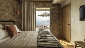 Une chambre avec un mur en pierre, un lit recouvert d’une couverture à rayures, une vue sur la mer et une porte en bois.