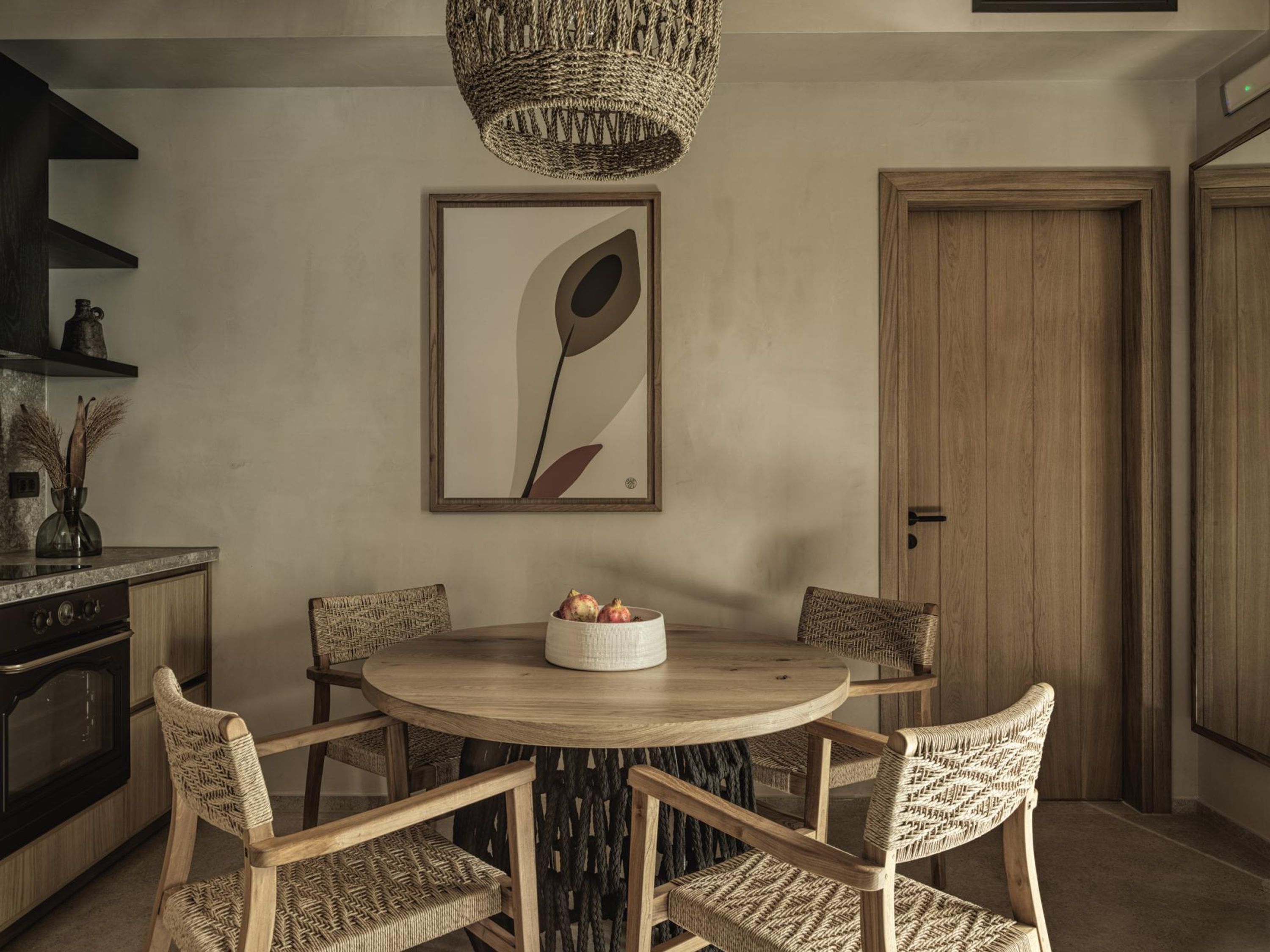 A dining area with a round wooden table, four wicker chairs, a woven pendant light, and a framed abstract painting on the wall.