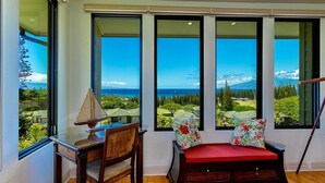 A room with a desk, chair, and a red cushioned bench with floral pillows, large windows with a view of a golf course and ocean. - Kapalua Golf Villas by Coldwell Banker Island Vacations (Lahaina)
