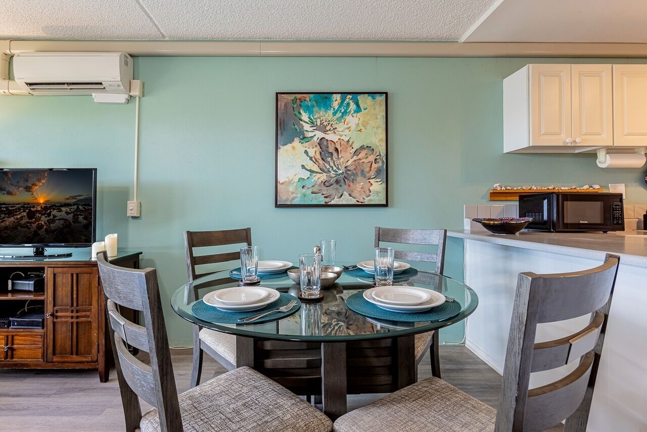 A dining area with a round glass table set for four, surrounded by wooden chairs. A painting hangs on the wall above the table.