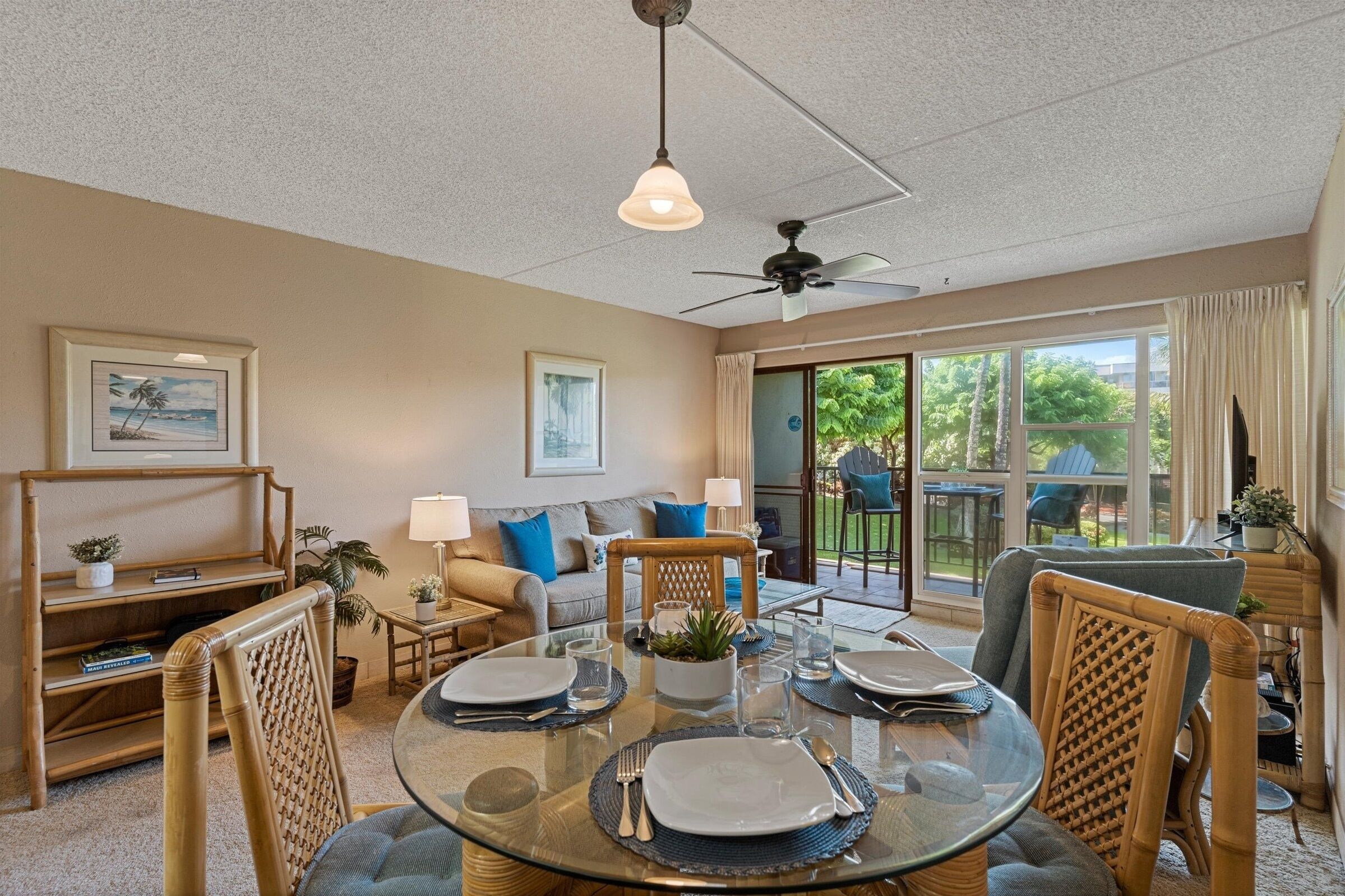 A dining area with a glass table set for four, surrounded by wicker chairs. There is a ceiling fan above and a large window with curtains to the right.
