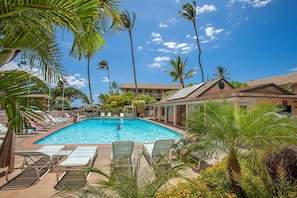 A swimming pool surrounded by lounge chairs and palm trees, with a building in the background. - Kihei Kai Nani #108 1 Bedroom Condo (Kihei)