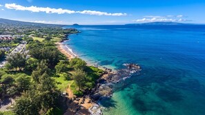 An aerial view of a coastal area with clear blue waters, a sandy beach, and a rocky shoreline.
