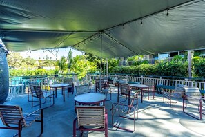 An outdoor patio area with tables and chairs, a covered section, and a view of greenery and buildings in the background. - Kihei Kai Nani #108 1 Bedroom Condo (Kihei)