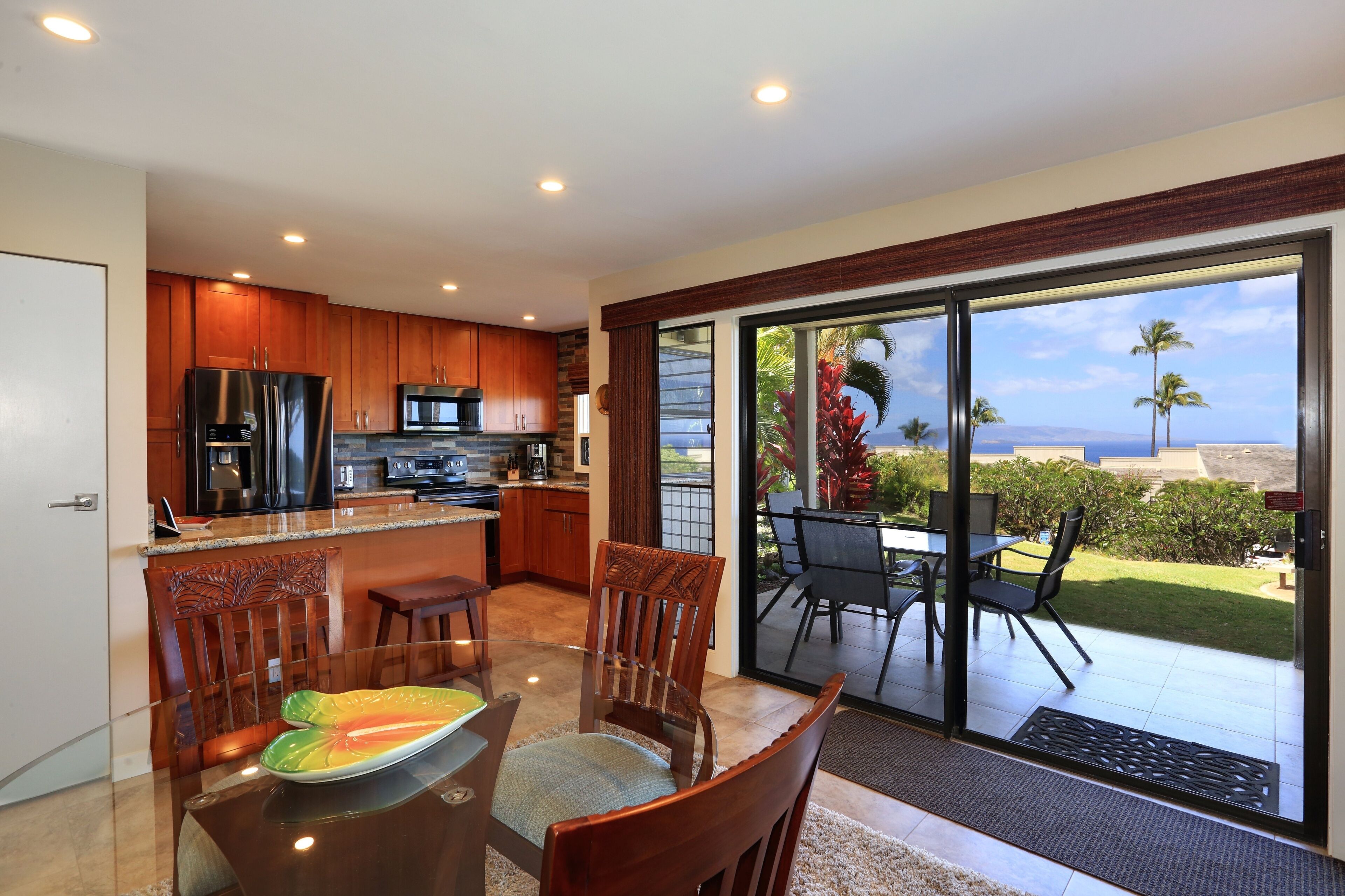 A modern kitchen with wooden cabinets and stainless steel appliances, a dining area with a glass table and chairs, and a sliding glass door leading to a patio with a view of the ocean.