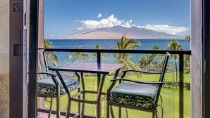 A balcony with a table and chairs, overlooking a coastal view with palm trees and a mountain. - Kihei Surfside, #609 1 Bedroom Condo by RedAwning (Kihei)