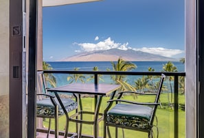 A balcony with a table and chairs, overlooking a coastal view with palm trees and a mountain. - Kihei Surfside, #609 1 Bedroom Condo by RedAwning (Kihei)