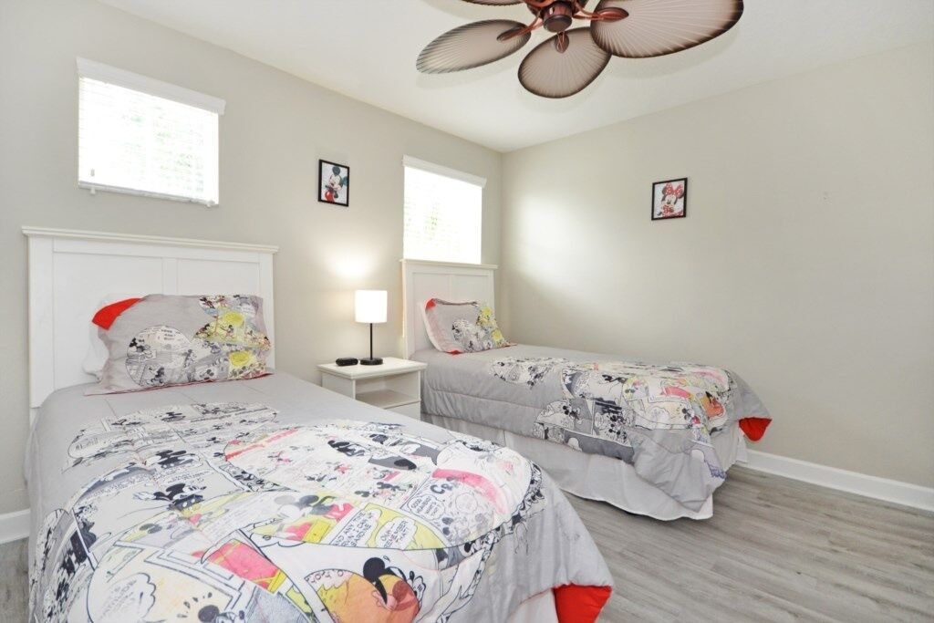 Une chambre avec deux lits, chacun recouvert d’une couverture à motifs de bandes dessinées, dotée d’un ventilateur de plafond et d’une table de chevet avec une lampe.