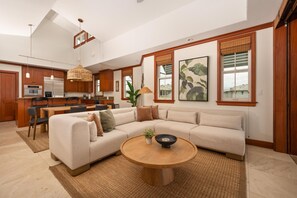 A spacious living area with a large L-shaped sectional sofa, a round wooden coffee table, and a kitchen in the background. - Big Island Kolea by Coldwell Banker Island Vacations (Waikoloa)
