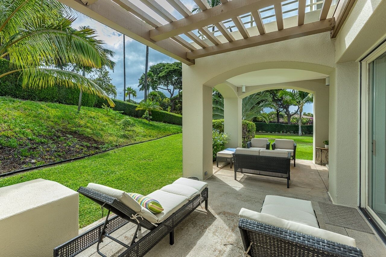 Une terrasse extérieure couverte, dotée de chaises longues et d’une table, qui offre une vue sur une pelouse bien entretenue et des palmiers.