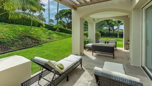 A covered outdoor patio with lounge chairs and a table, overlooking a well-maintained lawn and palm trees. - Palms at Wailea One Bedrooms by Coldwell Banker Island Vacations (Kihei)