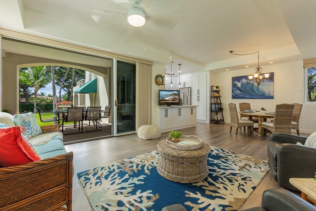 A spacious living area with a large sliding glass door leading to a patio, a dining area with wicker furniture, and a kitchen with modern appliances.