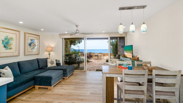 A modern living room with a dark blue sectional sofa, a wooden dining table with chairs, and a view of the beach through glass doors.