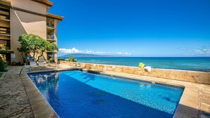 A swimming pool with a view of the ocean, surrounded by a building with multiple balconies and potted plants.
