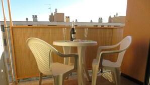 En liten balkong utomhus med två vita plaststolar och ett runt bord, en flaska champagne och ett glas på bordet.