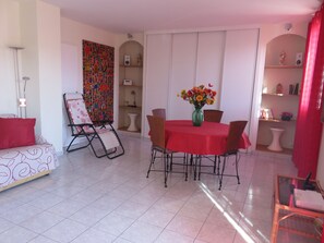 A dining area with a red table, chairs, and a vase of flowers. There is a sofa with a red pillow, a chair with a patterned cushion, and a white floor.