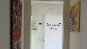En hall med en dörr som leder in till ett rum. På dörren finns en skylt med texten ”Ha en bra dag!” tillsammans med ett tassavtryck och en symbol som visar att rökning är förbjuden. På den vänstra väggen hänger en färgstark, abstrakt målning och på den högra väggen en mindre målning.