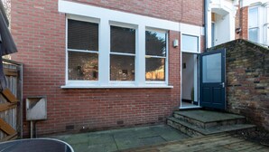 A red brick house with a white-framed window, a blue door, and a wooden deck. - Serene and Spacious 1 Bedroom Garden Flat in Clapton (London)