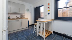 A kitchen with white cabinets, a wooden stool, a window with a view, and a clock on the wall. - Serene and Spacious 1 Bedroom Garden Flat in Clapton (London)