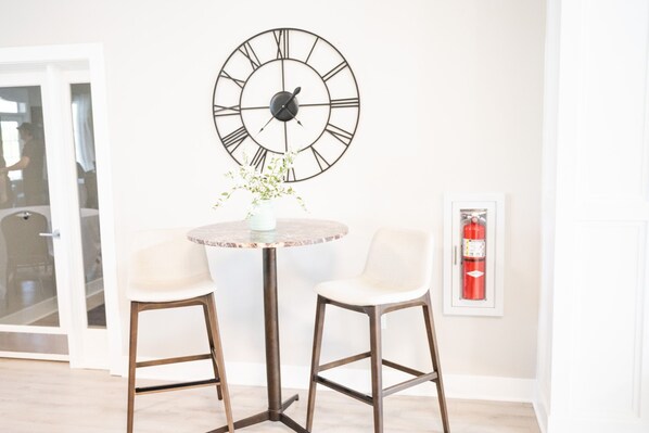 A room with a round table, two chairs, a wall clock, a small vase with flowers, and a fire extinguisher.