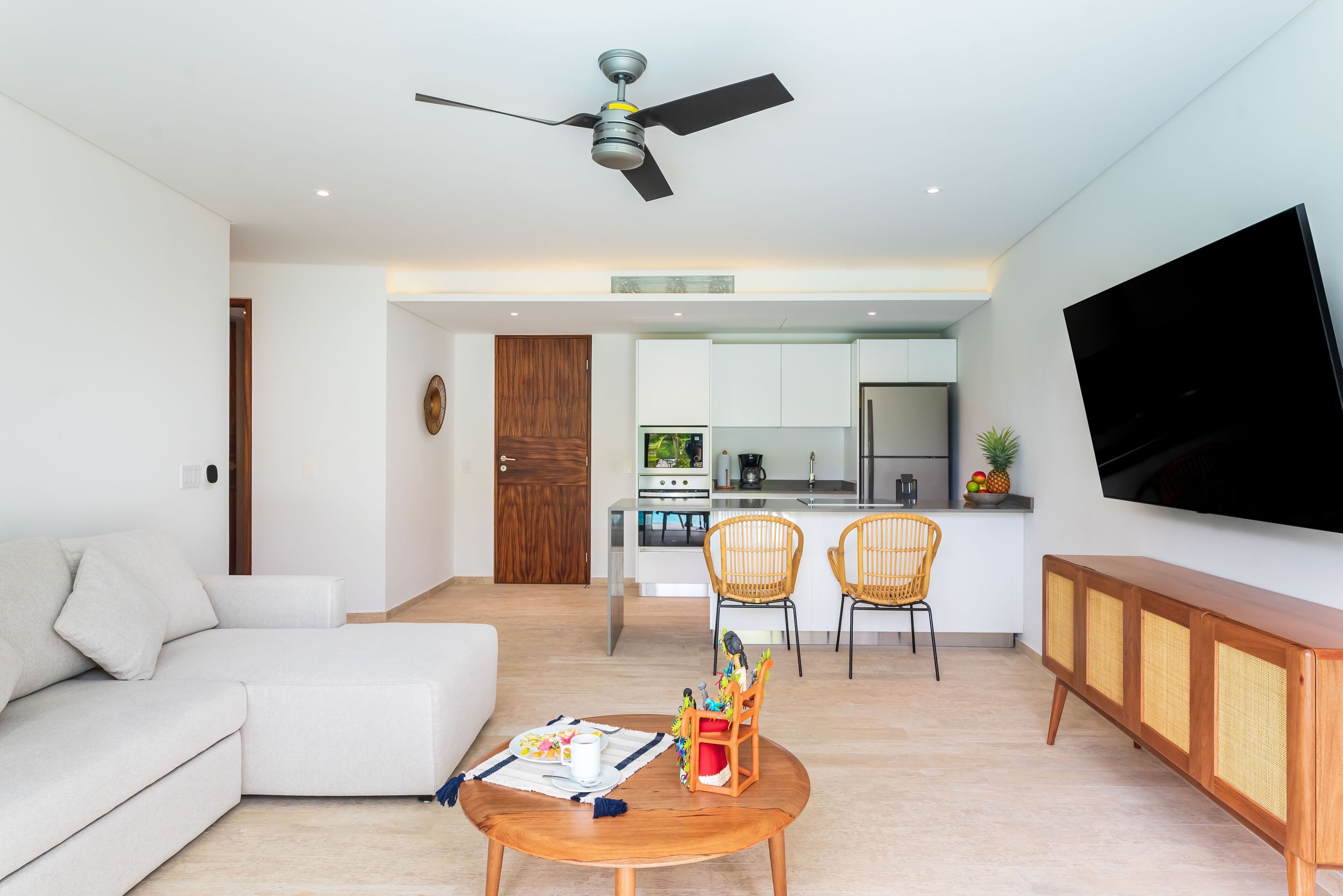 A modern living room with a sofa, a coffee table, and a TV. There is a kitchen in the background with a dining area.