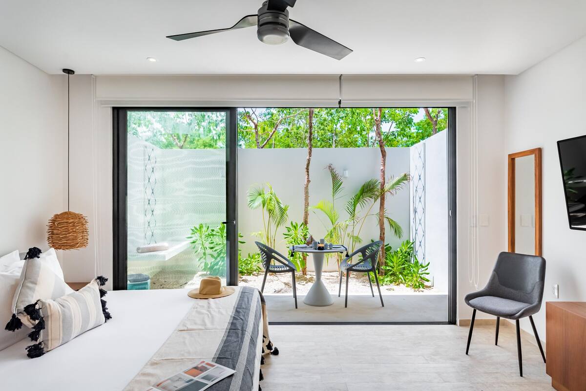 A modern bedroom with a bed, a ceiling fan, a dining area with chairs, and a view of greenery outside.