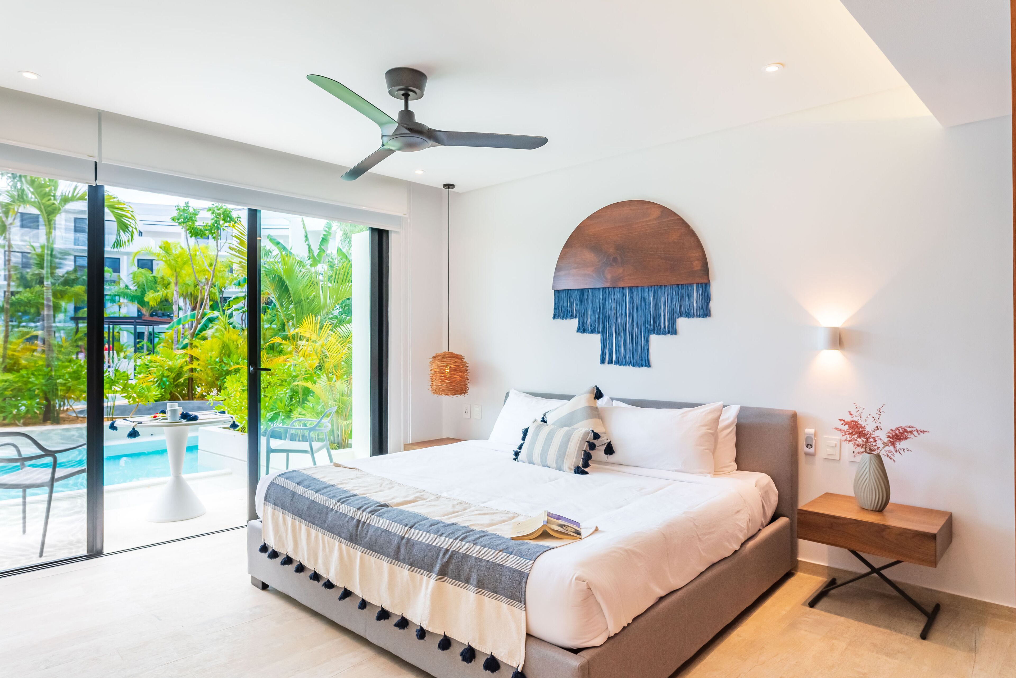 Une chambre moderne avec un grand lit, un ventilateur de plafond, une vue sur une piscine et une table de chevet en bois sur laquelle est posé un vase.