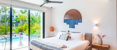 Une chambre moderne avec un grand lit, un ventilateur de plafond, une vue sur une piscine et une table de chevet en bois sur laquelle est posé un vase.