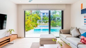 Una sala moderna con televisor grande, sofá, una mesita de centro y vistas a una piscina y vegetación.