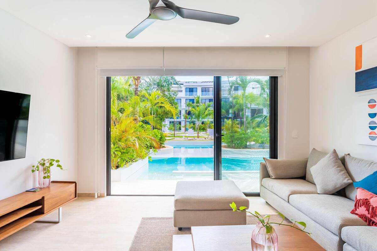 A modern living room with a large TV, a sofa, a coffee table, and a view of a swimming pool and greenery outside.