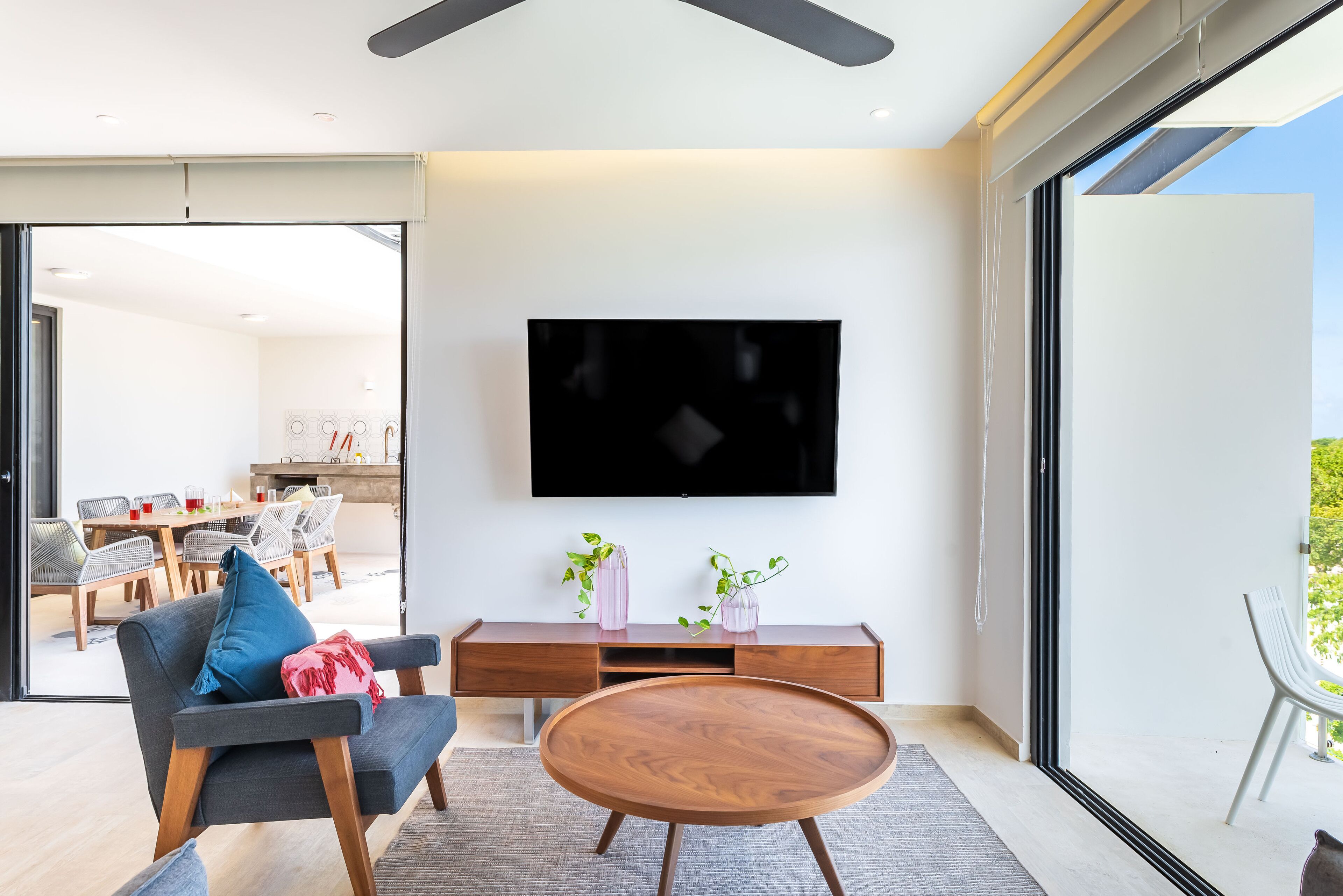 A modern living room with a flat-screen TV, a sofa, a coffee table, and a dining area visible through a glass door.