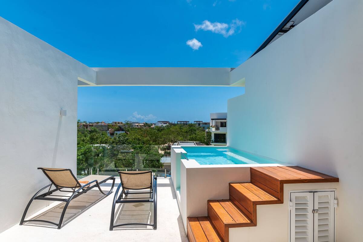 A rooftop terrace with a swimming pool, two lounge chairs, wooden steps, and a view of the city.