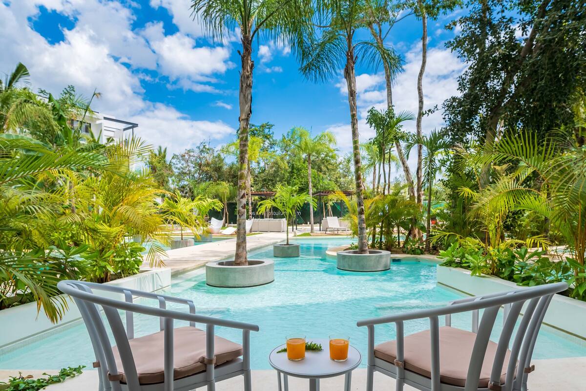 A poolside area with lounge chairs and a table, surrounded by palm trees and greenery.