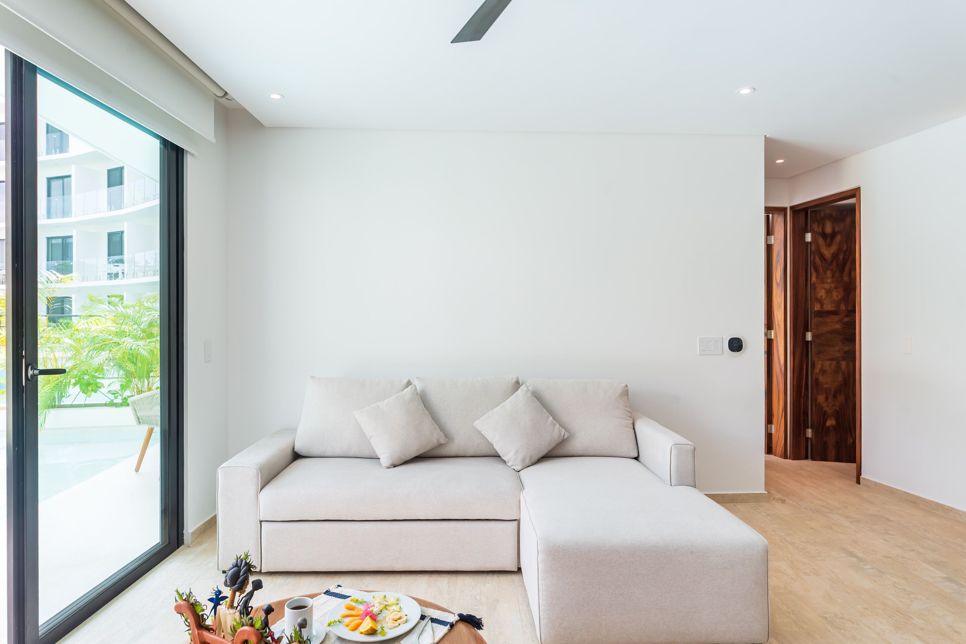 A modern living room with a light-colored sectional sofa, a coffee table with a plate of food, and a large window offering a view of a building and greenery outside.