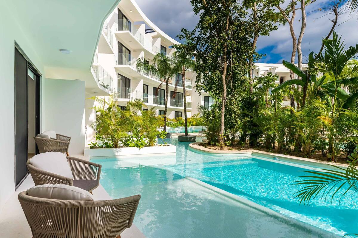 A modern hotel with a swimming pool surrounded by lush greenery and a white building with balconies.