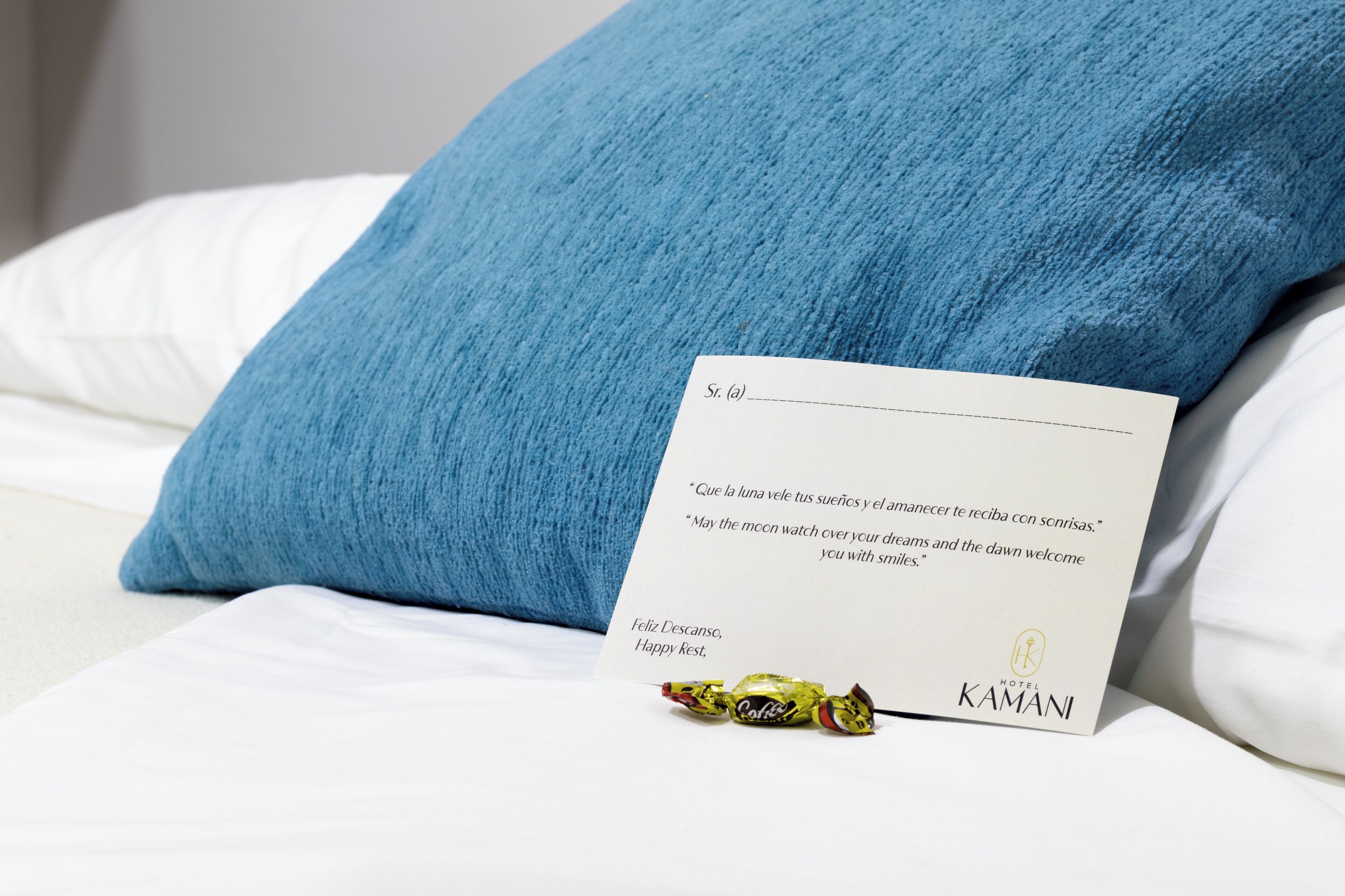 Una habitación de hotel con una cama blanca, un almohadón azul, una tarjeta con una cita y un regalo envuelto.