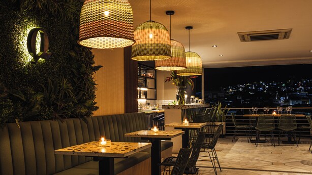 A restaurant with a green cushioned bench, tables with candles, wicker pendant lights, and a city view at night.