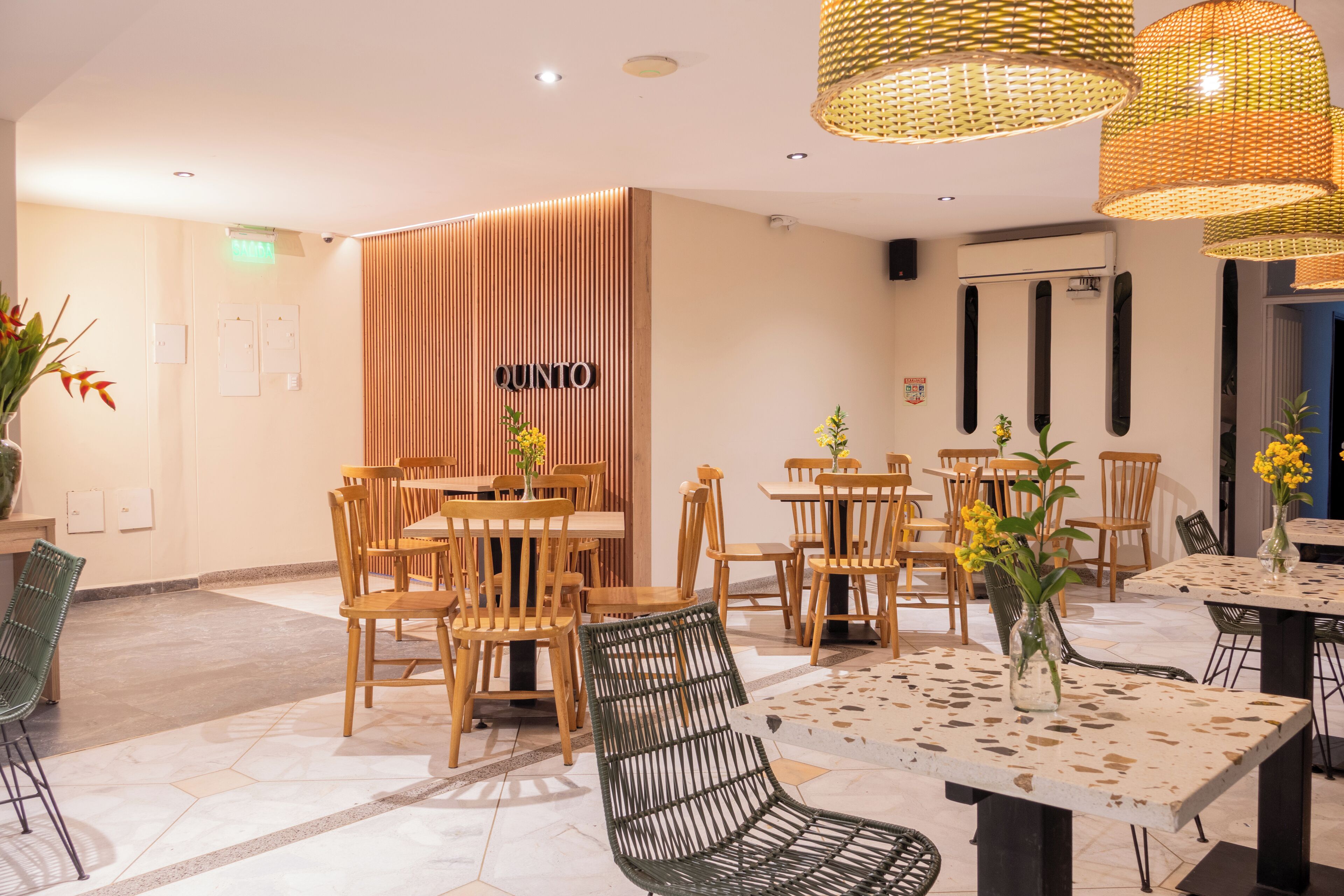 A restaurant interior with wooden tables and chairs, a woven ceiling light fixture, and a wall-mounted air conditioning unit.
