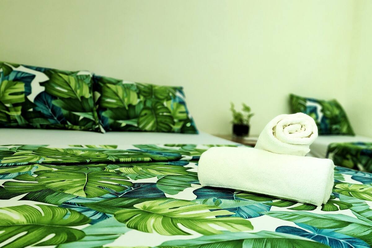 A hotel room with a bed featuring a tropical leaf pattern bedspread, a neatly rolled white towel, and a small potted plant on the nightstand.