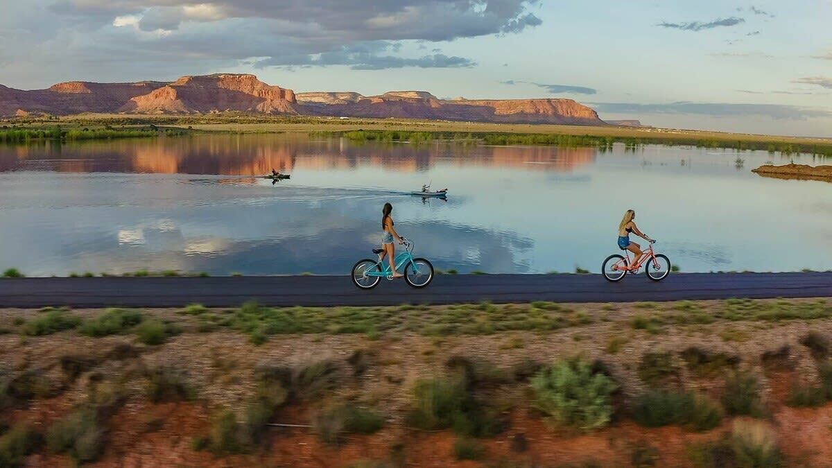 Un lac paisible, avec deux cyclistes sur une route au premier plan, le tout entouré d’un paysage naturel et de montagnes lointaines.