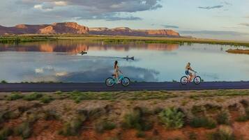 Un lac paisible, avec deux cyclistes sur une route au premier plan, le tout entouré d’un paysage naturel et de montagnes lointaines.