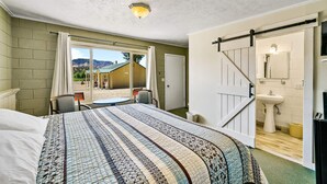 A bedroom with a large bed, a sliding door to a bathroom, a view of the outdoors, and a small table with chairs. - Mt Carmel Motel close to Zion National Park (Orderville)