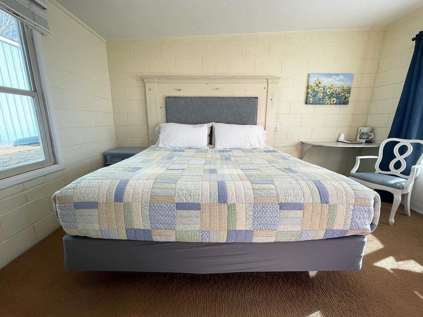 A single-bed room with a quilted bedspread, a grey headboard, a small bedside table with a lamp, a chair, and a painting on the wall.