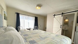 Un dormitorio con una cama grande, una mesita, dos sillas, una ventana con cortinas y un baño con lavamanos y espejo.