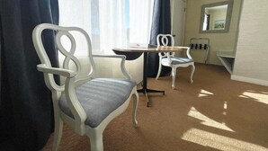 Una habitación con una silla blanca con cojín estampado, una pequeña mesa redonda y un ventanal con cortinas.