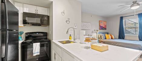 A modern kitchen with a refrigerator, stove, microwave, and sink. There is a dining area with a table and chairs, and a bed with a view of the outside.