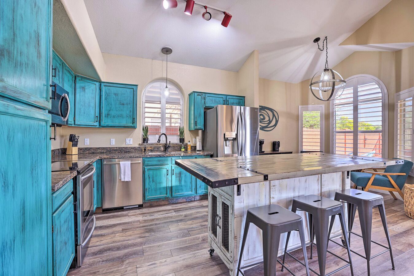 A kitchen with teal cabinets, stainless steel appliances, a central island with stools, and large windows.